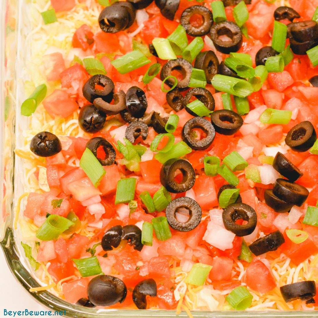 Spicy ranch 7-layer taco dip is Layers of taco-seasoned refried beans, guacamole, sour cream with ranch, lettuce, cheese, pico de gallo, olive, and cheese!