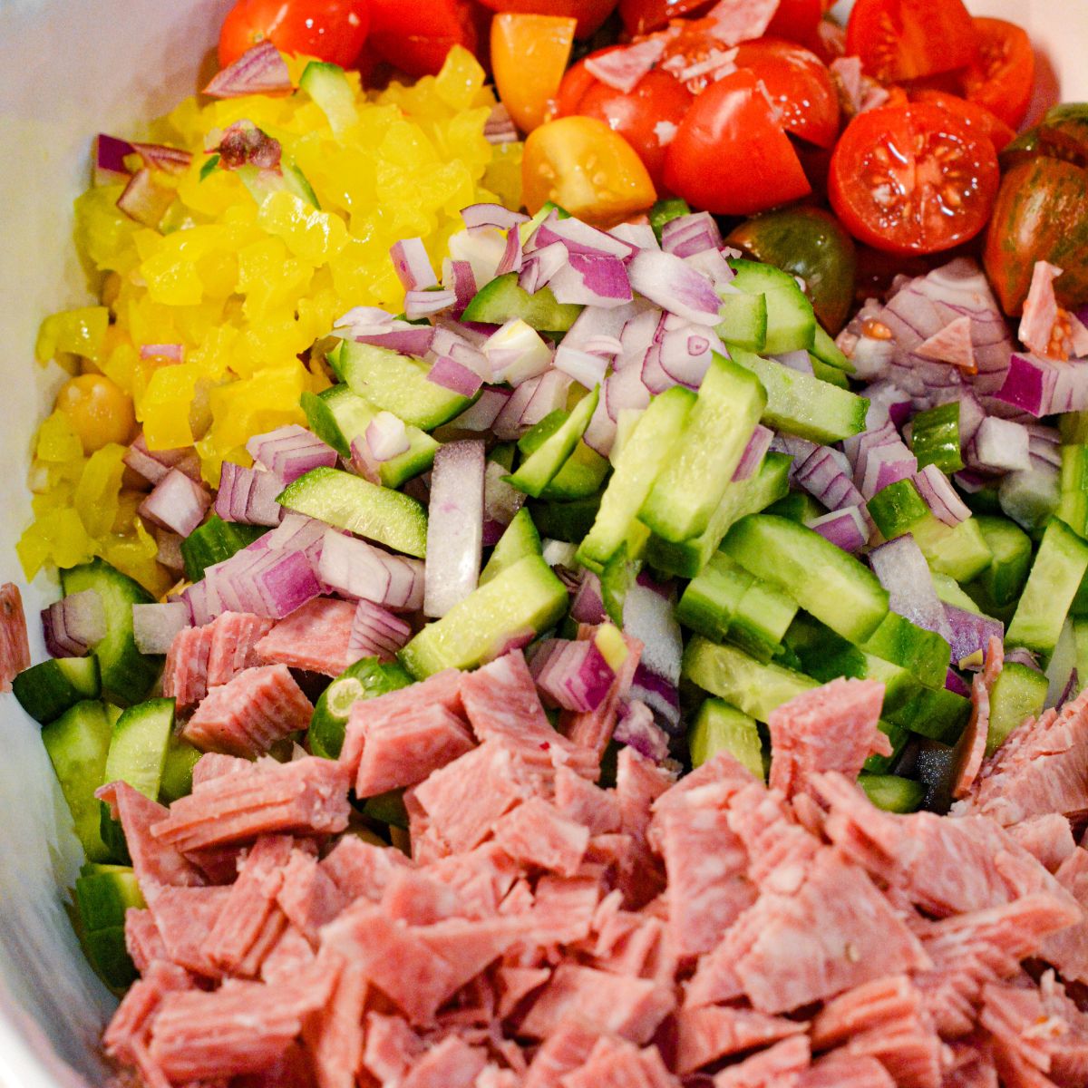 To the bowl, add the sliced red onion, banana peppers, salami, black olives, and cherry tomatoes. 