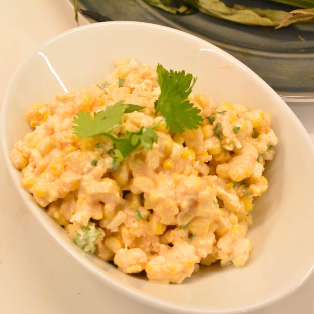Bowl of creamy street corn salad garnished with cilantro next to grilled corn cobs