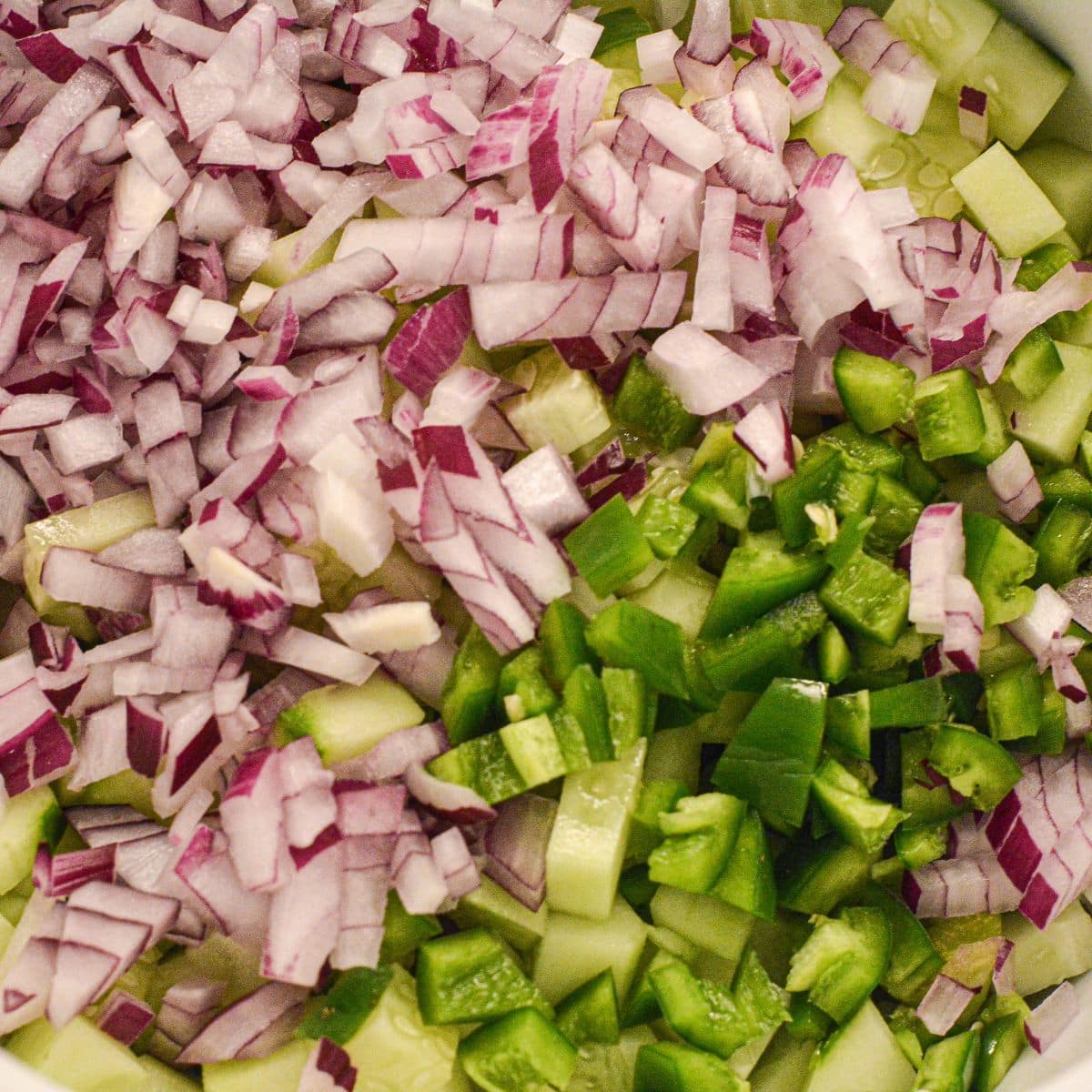Diced red onion and jalapeño on top of chopped cucumber in a mixing bowl